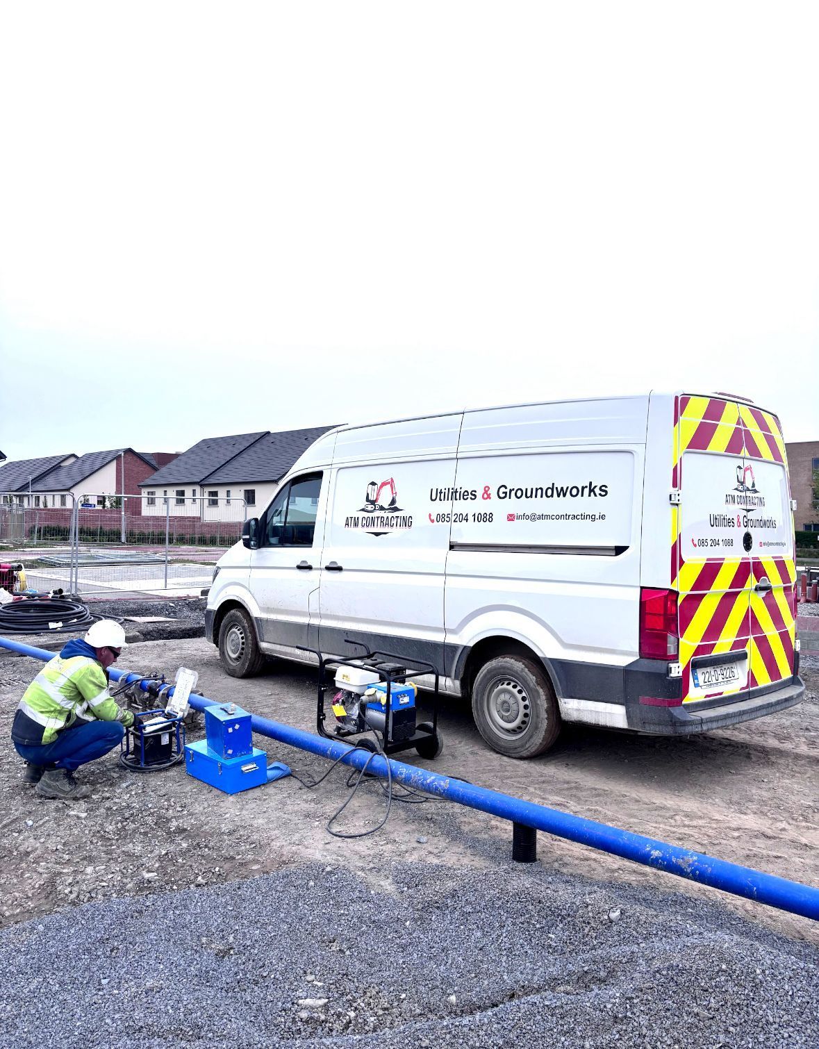 Construction worker near white van, connecting blue pipes. Gray gravel, buildings, overcast sky.