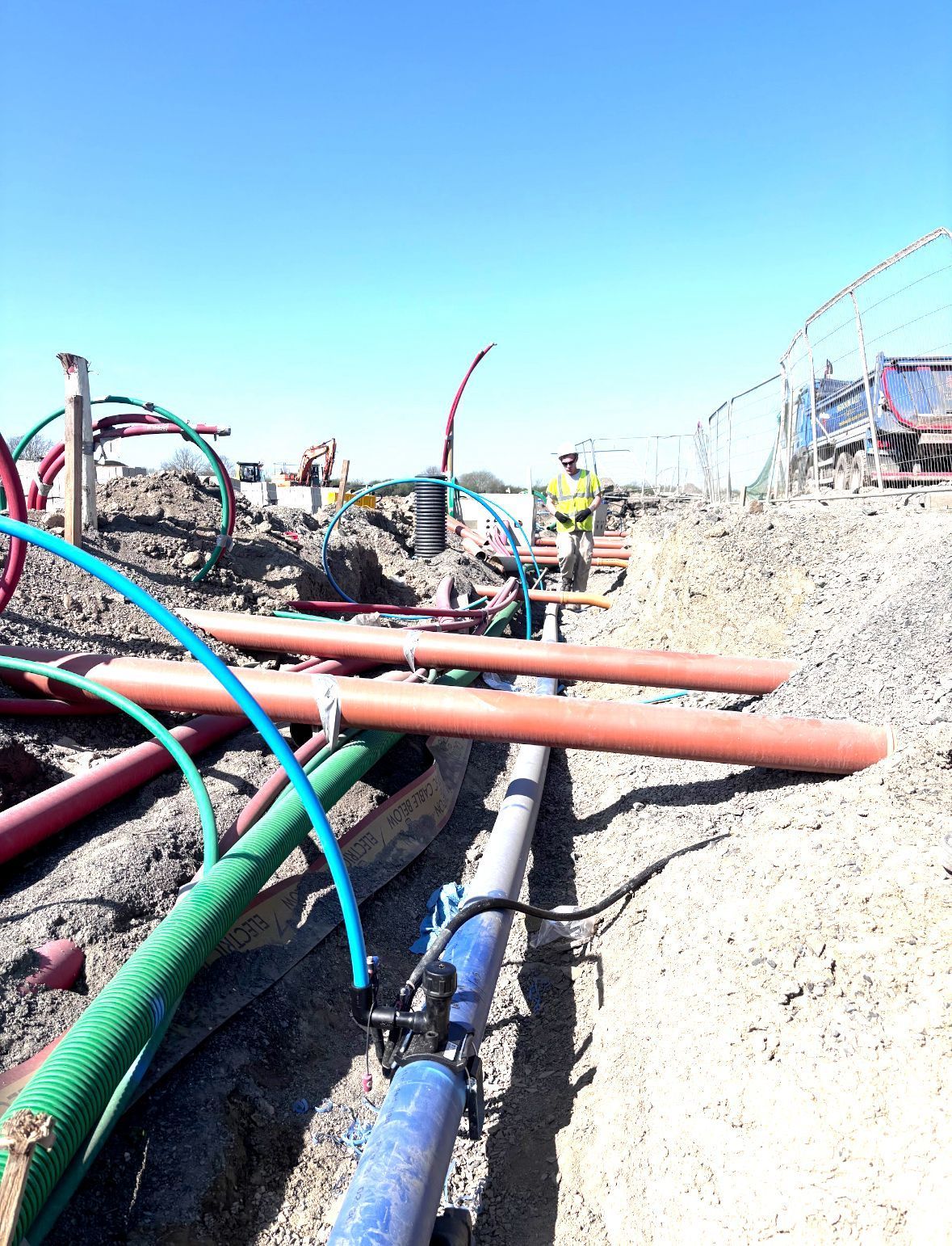 Construction site with buried pipes of various colors; a worker in high-vis vest stands in background.
