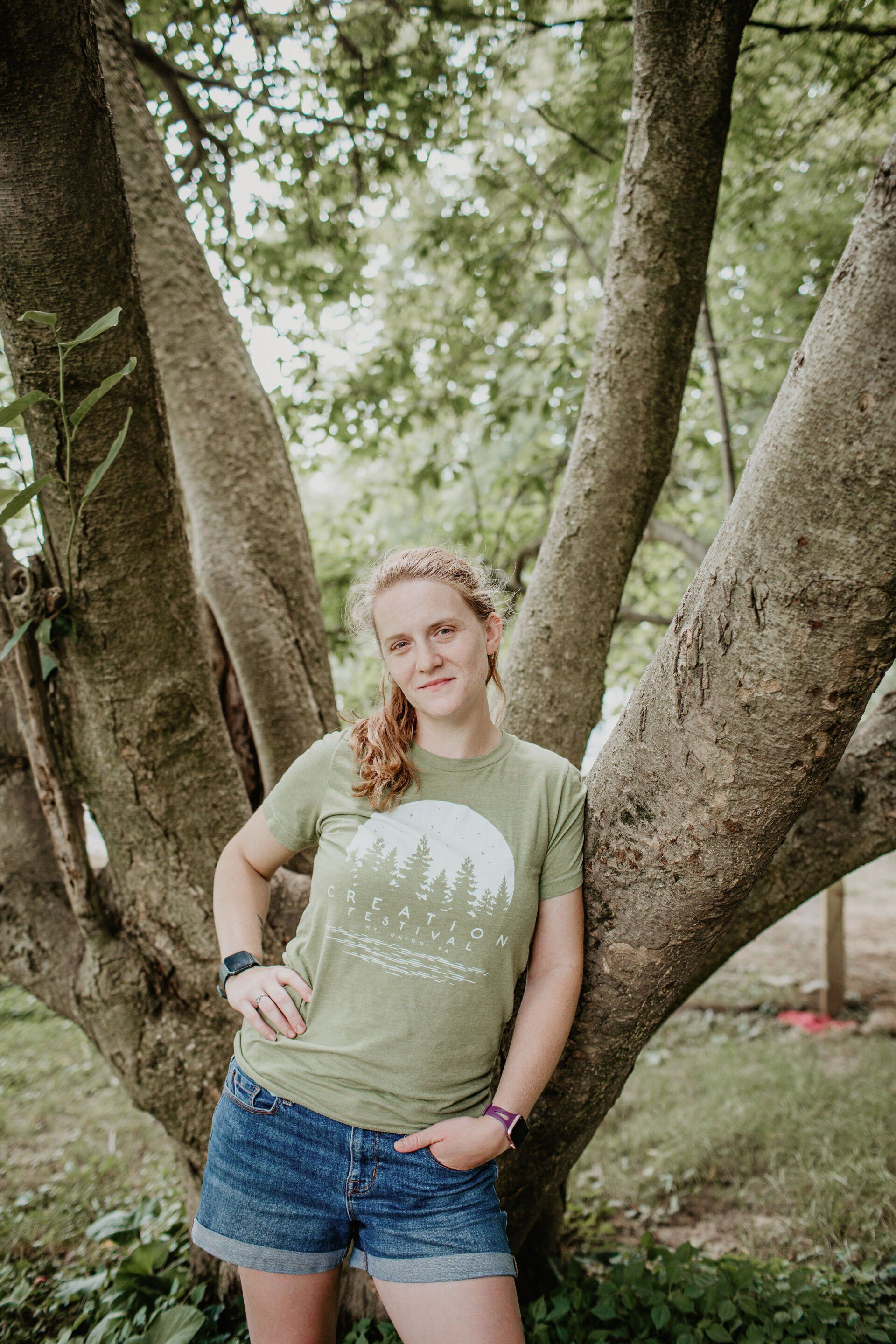 A woman in a green t-shirt and blue shorts is leaning against a tree.