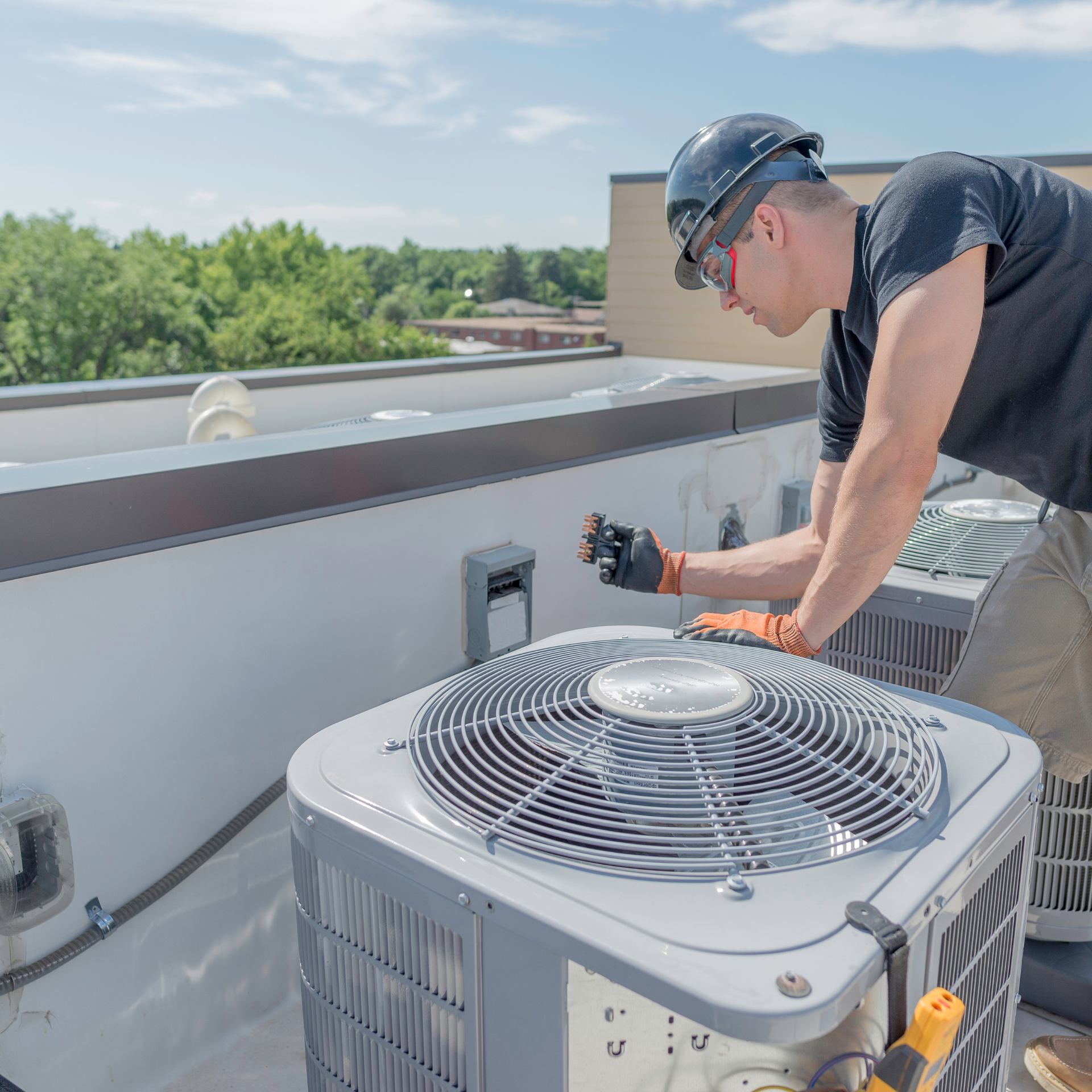 HVAC technician in a hard hat and safety glasses on a rooftop, working on an air conditioning unit.