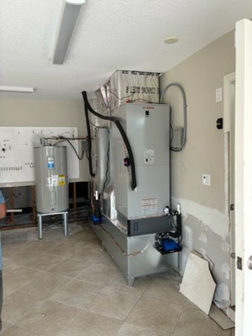 A room with HVAC equipment; large, gray tanks, black pipes, and a smaller water heater stand on a tiled floor.