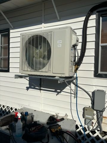 Outdoor AC unit mounted on a white house, with visible wiring and a shelf.