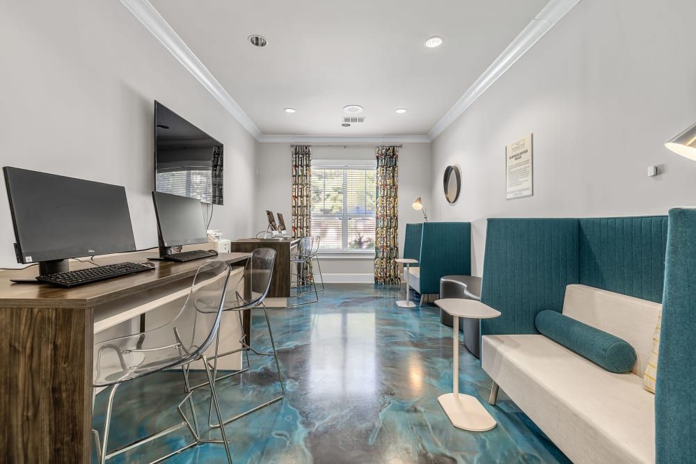 A modern clubhouse interior featuring a wooden desk with two desktop computers, clear acrylic chairs, and teal upholstered bench seating. The room has a glossy, marbled blue floor, white walls with decorative curtains, and recessed ceiling lights. A framed certificate and a round mirror hang on the wall, creating a professional ambiance at Marq Perimeter in Atlanta, GA.