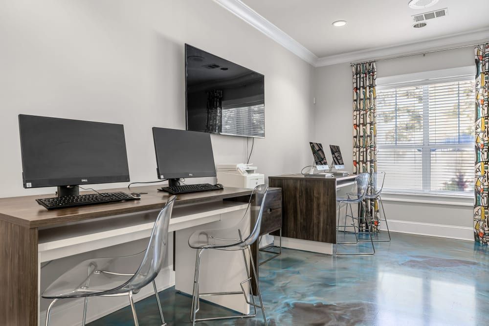 A modern business center with two workstations setup side by side. Each station has a desktop computer with a black monitor on a wooden desk, accompanied by a clear acrylic chair. To the right, a window with blinds is partially covered by colorful patterned drapes. The floor has a glossy, blue-tinted finish that reflects the room's elements, adding a unique touch to the space at Marq Perimeter in Atlanta, GA.