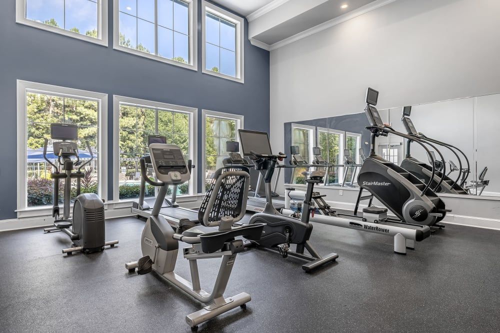 A modern and well-lit gym with tall windows, grey walls, and a mix of cardio equipment including treadmills, a recumbent bike, elliptical machines, a StairMaster, and a WaterRower rowing machine. The space is clean and appears to be private or part of a small fitness center at Marq Perimeter in Atlanta, GA.