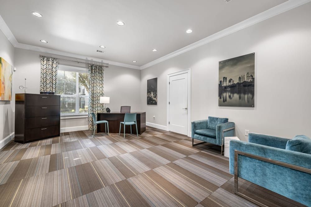 A modern and well-lit leasing office with multi-toned brown striped flooring. To the left, there's a dark wood dresser beside a workspace with a desk, a lamp, and two turquoise chairs in front of a window with patterned curtains. On the right, two plush teal armchairs sit facing each other with a side table in between. The walls are adorned with framed cityscape photographs, and a white door is visible on the right-hand side at Marq Perimeter in Atlanta, GA.