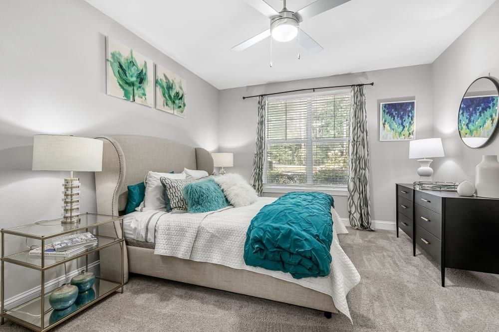 Modern bedroom with neutral walls, featuring a large bed with an upholstered headboard and teal blue accents in the bedding and wall art. Stylish black nightstands, decorative mirrors, and a ceiling fan complete the room's decor at Marq Perimeter in Atlanta, GA.