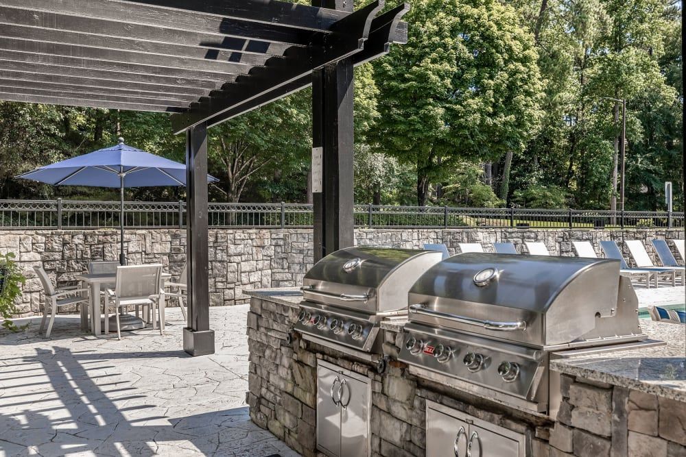 An outdoor poolside patio with two stainless steel barbeque grills under a pergola, near a dining area with a table, chairs, and a blue umbrella. Lounge chairs are lined up by the pool in the background, which is surrounded by a stone wall and lush green trees under a clear sky at Marq Perimeter in Atlanta, GA.