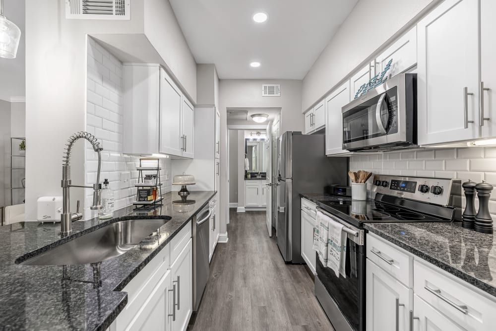 Modern kitchen interior with white cabinetry, stainless steel appliances, and granite countertops. The space features a double sink with a high-arc faucet, subway tile backsplash, and recessed lighting. A corridor is visible in the background, leading to another part of the home at Marq Perimeter in Atlanta, GA.