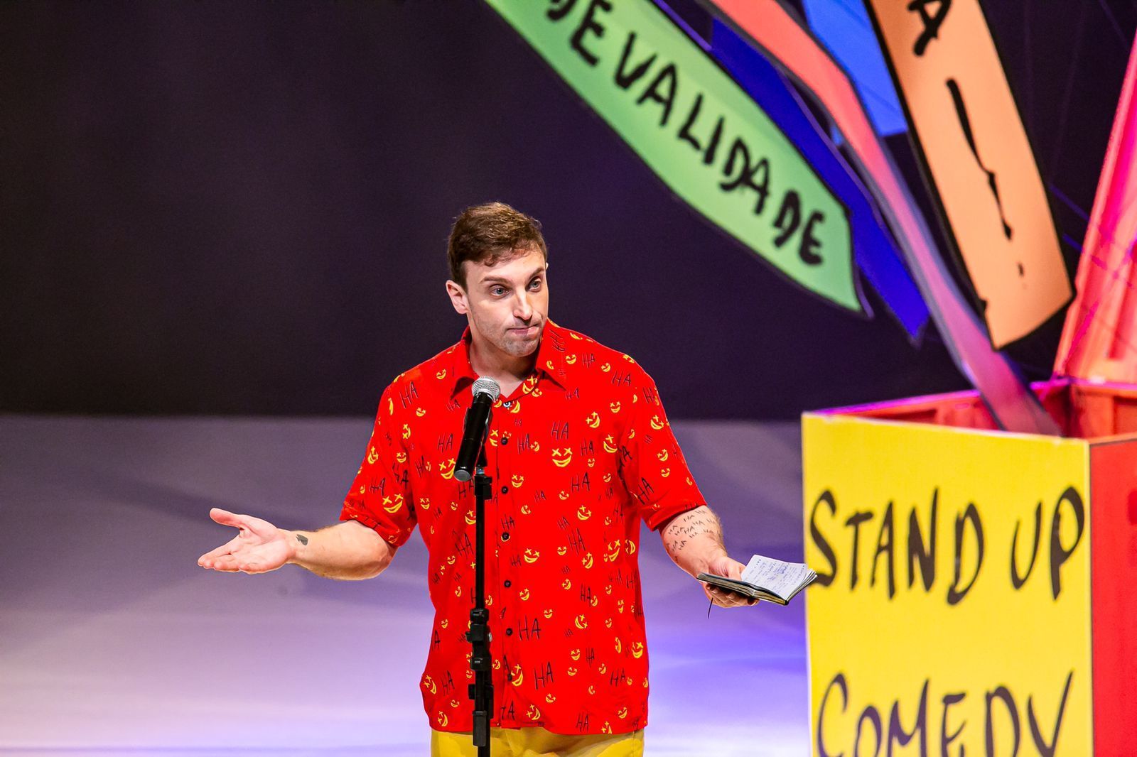 Um homem de camisa vermelha está parado em frente a um camarote de stand up comedy.