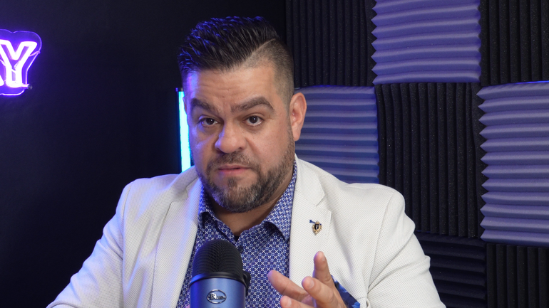 Um homem com barba está sentado em frente a um microfone em um estúdio de gravação.