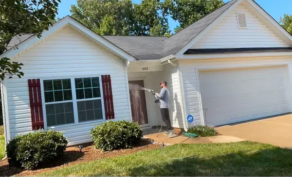 Man power washing a white house with a two-car garage. Red shutters flank a window. Green grass and blue sky.
