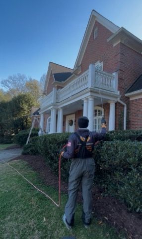 Person spraying the front of a red brick house with white columns. The person is wearing a backpack.