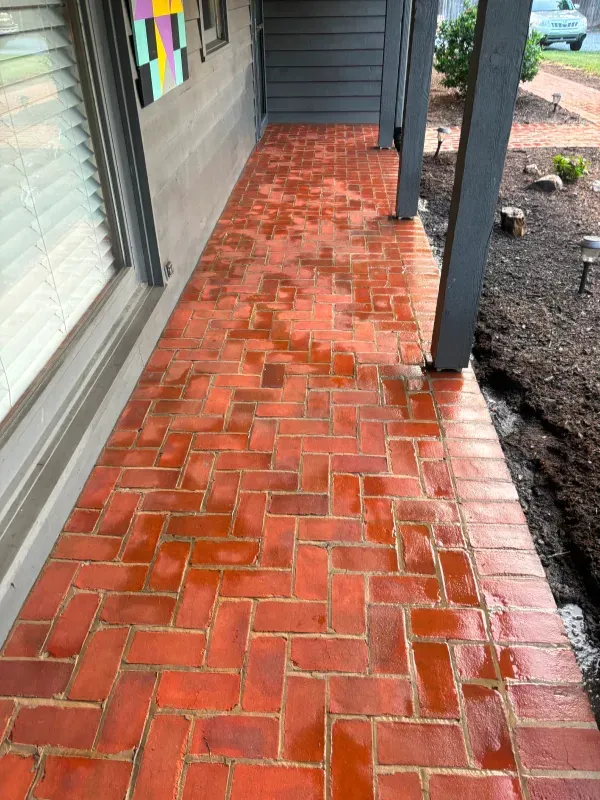 Red brick herringbone-patterned porch with wet surface. Gray posts and siding. Lawn and window to the left.