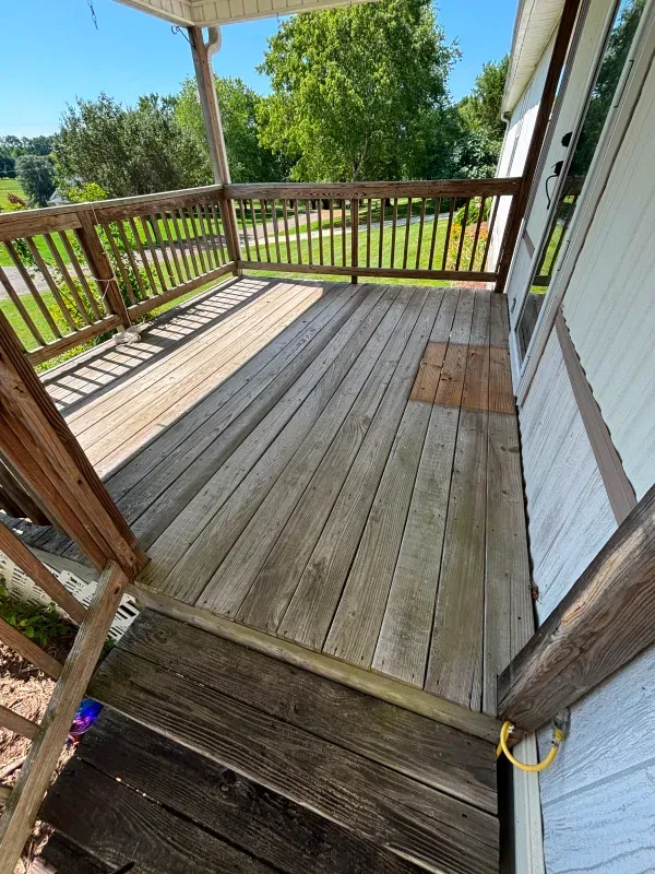 Wooden deck with weathered planks and railing, attached to a house. Bright, sunny day.