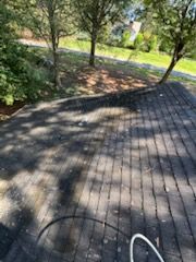Dark asphalt shingle roof with shadows and a nearby grassy area with trees in the background.