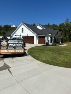 A white house with a dark roof and brown garage doors sits on a green lawn. Pressure washing truck parked in the driveway.