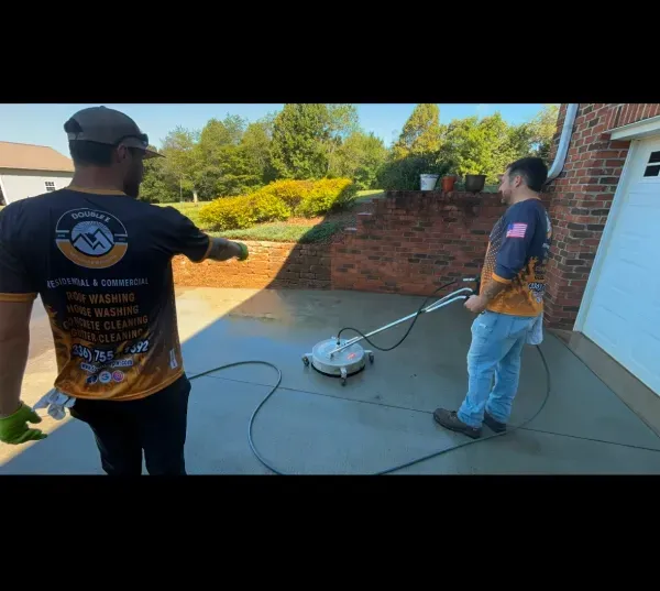 Two men pressure washing a concrete surface next to a brick building. One points; the other operates the washer.