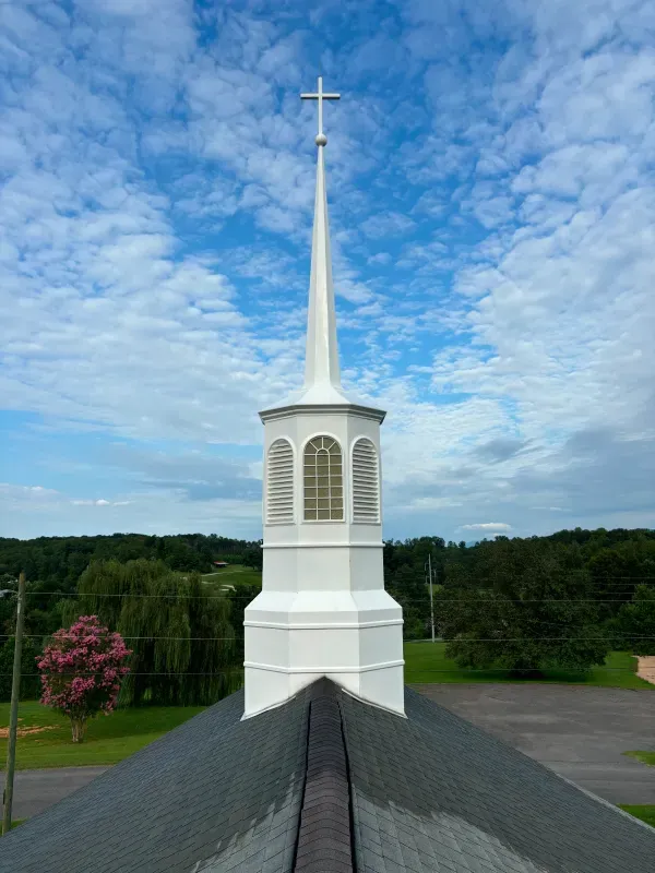 White church steeple with cross against a cloudy sky, seen from a high angle over a dark roof.