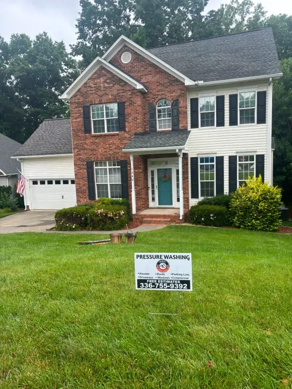 Two-story house with brick and white siding, black shutters, and green lawn. A sign in front.