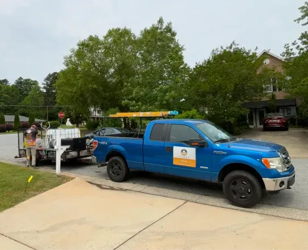 Blue truck with trailer parked on driveway. Person in reflective vest near the trailer. House in background.
