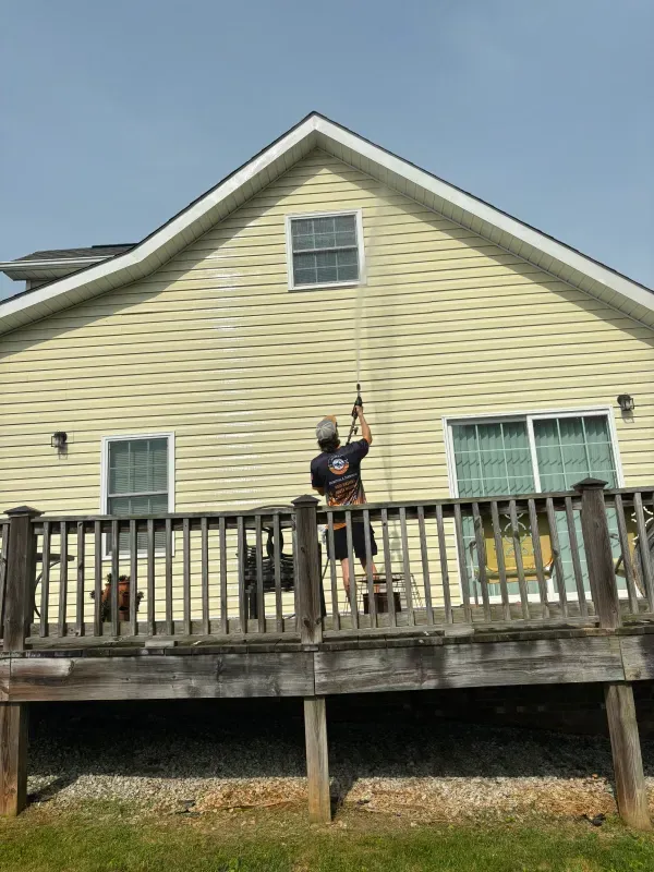 Man pressure washing a yellow house siding from a wooden deck on a sunny day.