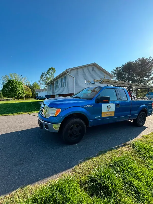 Blue pickup truck parked in front of a house on a sunny day. Truck has ladder on top and logo on the side.