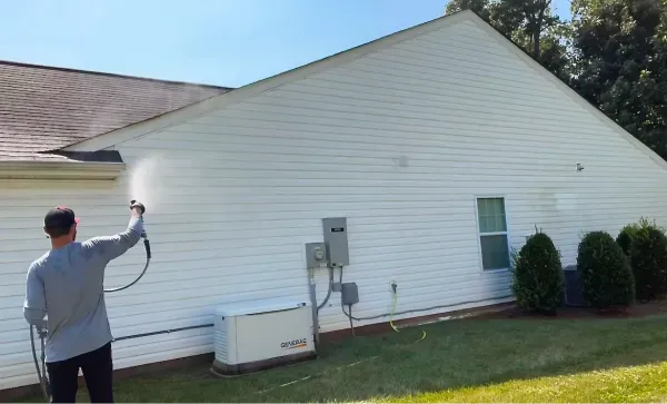 Person pressure washing the white siding of a house with a green lawn.