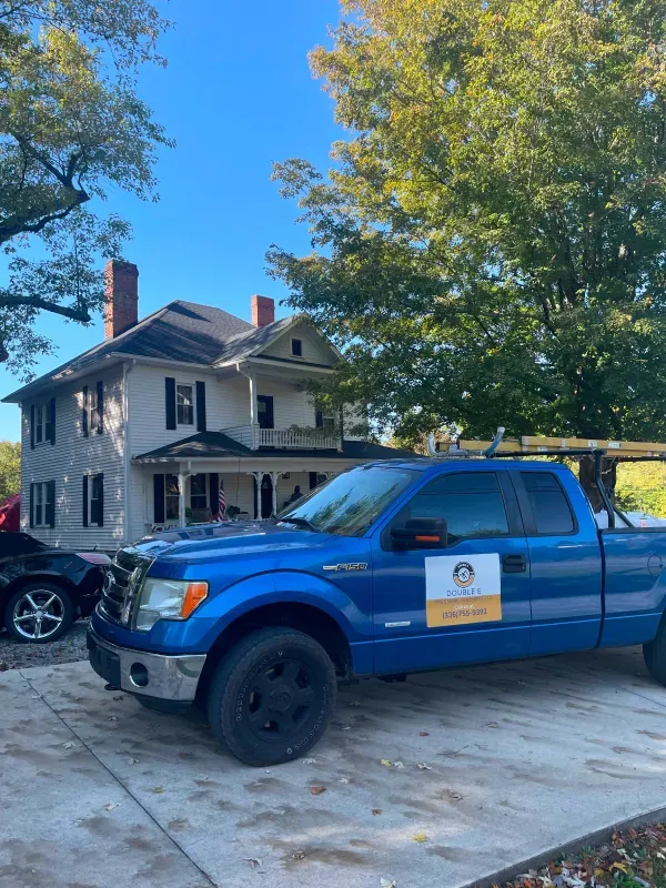 Blue pickup truck parked in front of a two-story white house with black shutters, trees, and blue sky.