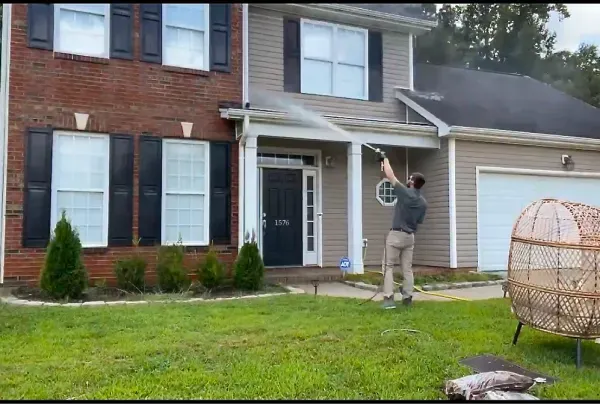 Man power washing the exterior of a two-story house with brick and siding.