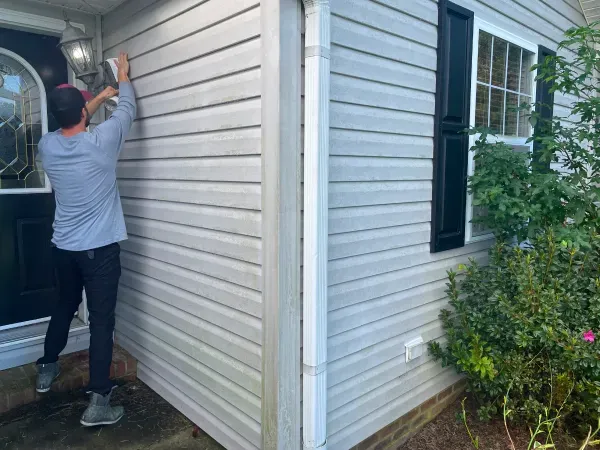 Man working on light fixture on house's siding. Gray siding, black door and shutters.