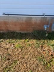 A brown foundation wall with a black pipe, gas meter, and overgrown grass in a yard.