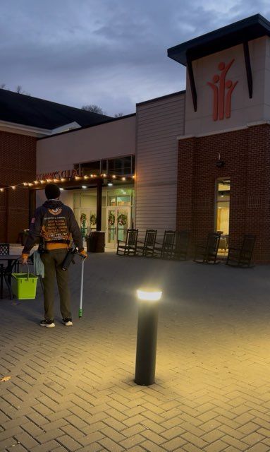 Person holding supplies walks toward a building with a red logo. Evening scene. Brick walkway.