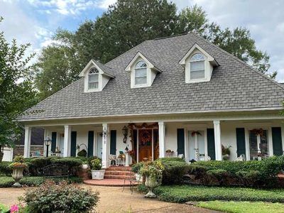 Two-story white house with a gray roof and three dormer windows; a porch with green shutters and manicured landscaping.