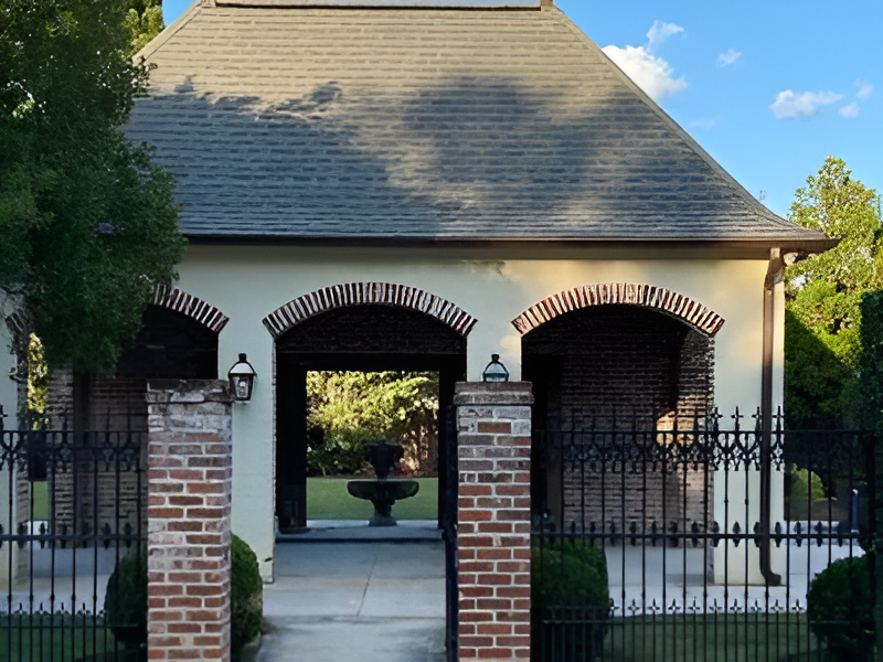 Brick arched building with black wrought-iron fence, facing a fountain in the garden.