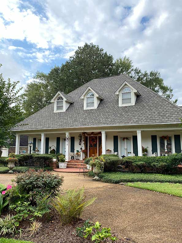 A two-story home with a white porch and dark green shutters, surrounded by green shrubs and a winding path.