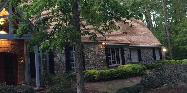 Stone cottage with brown roof and black shutters, surrounded by trees.