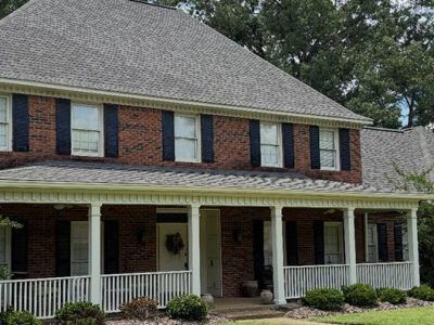 Two-story brick house with gray roof, white porch and trim, black shutters, and green bushes.