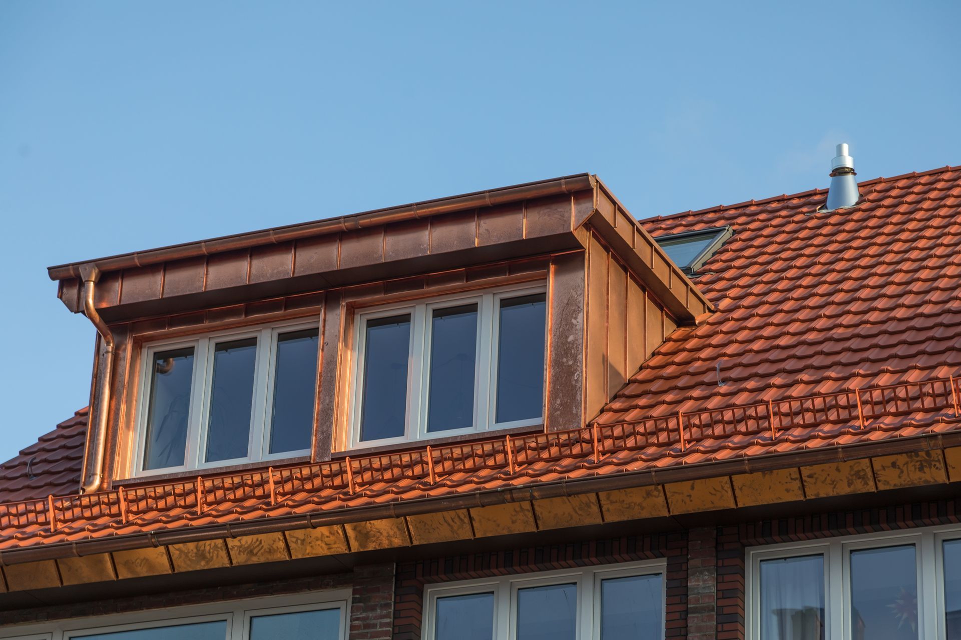 Copper-trimmed dormer with windows on a red tile roof against a blue sky.