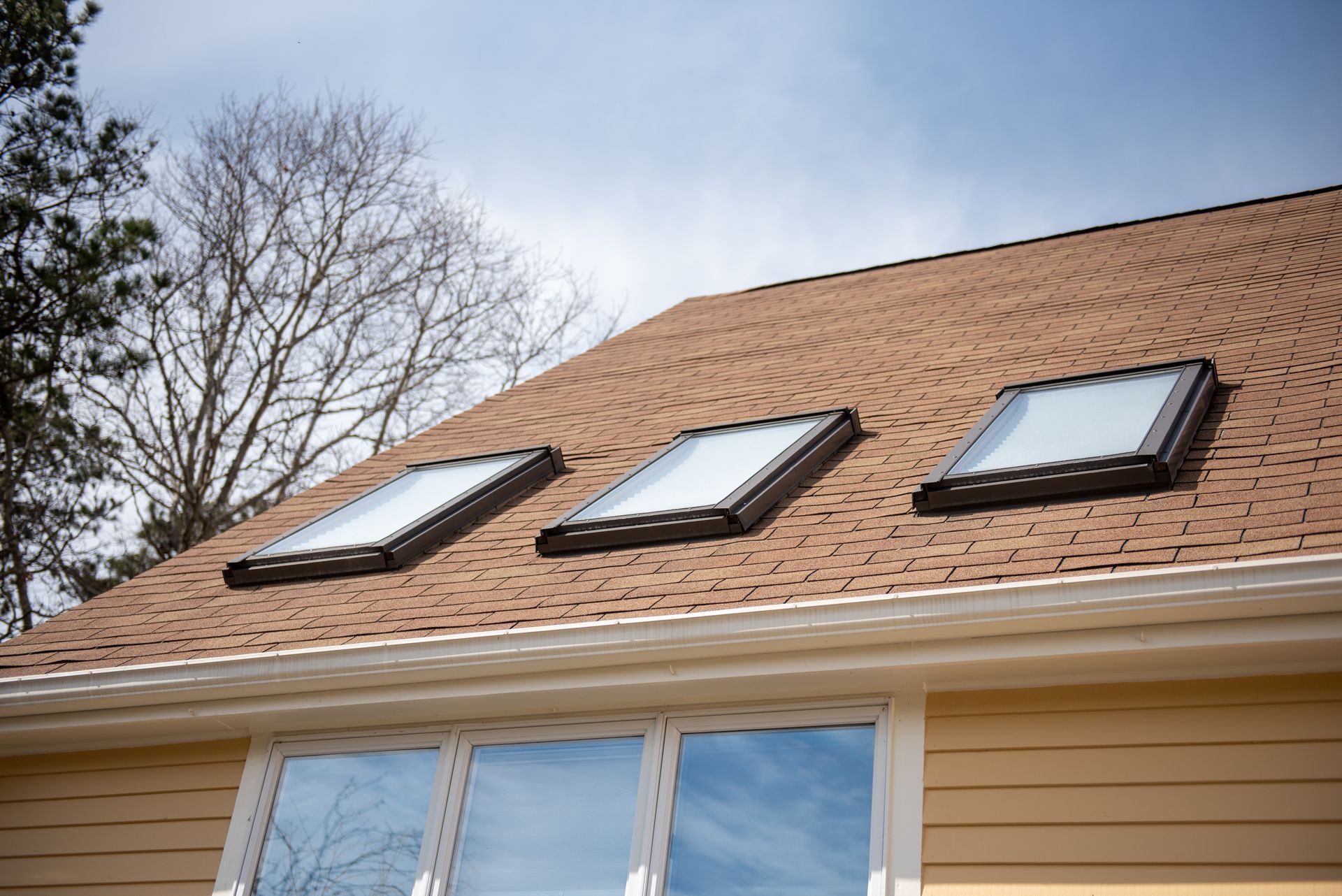 Three skylights on a brown shingled roof, above a yellow house with white trim and a cloudy sky.