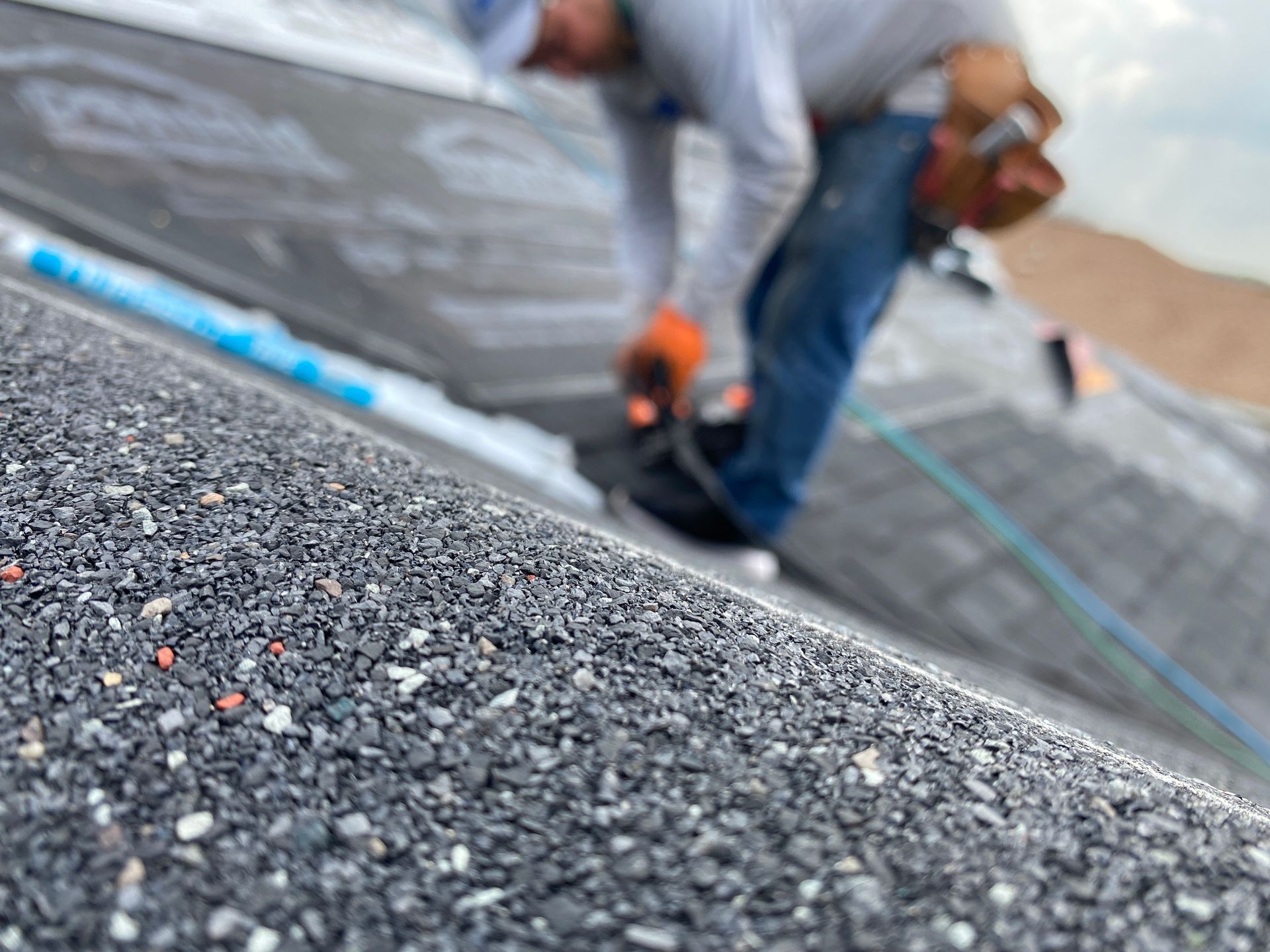 A worker in blue jeans and an orange glove installs gray asphalt shingles on a roof.