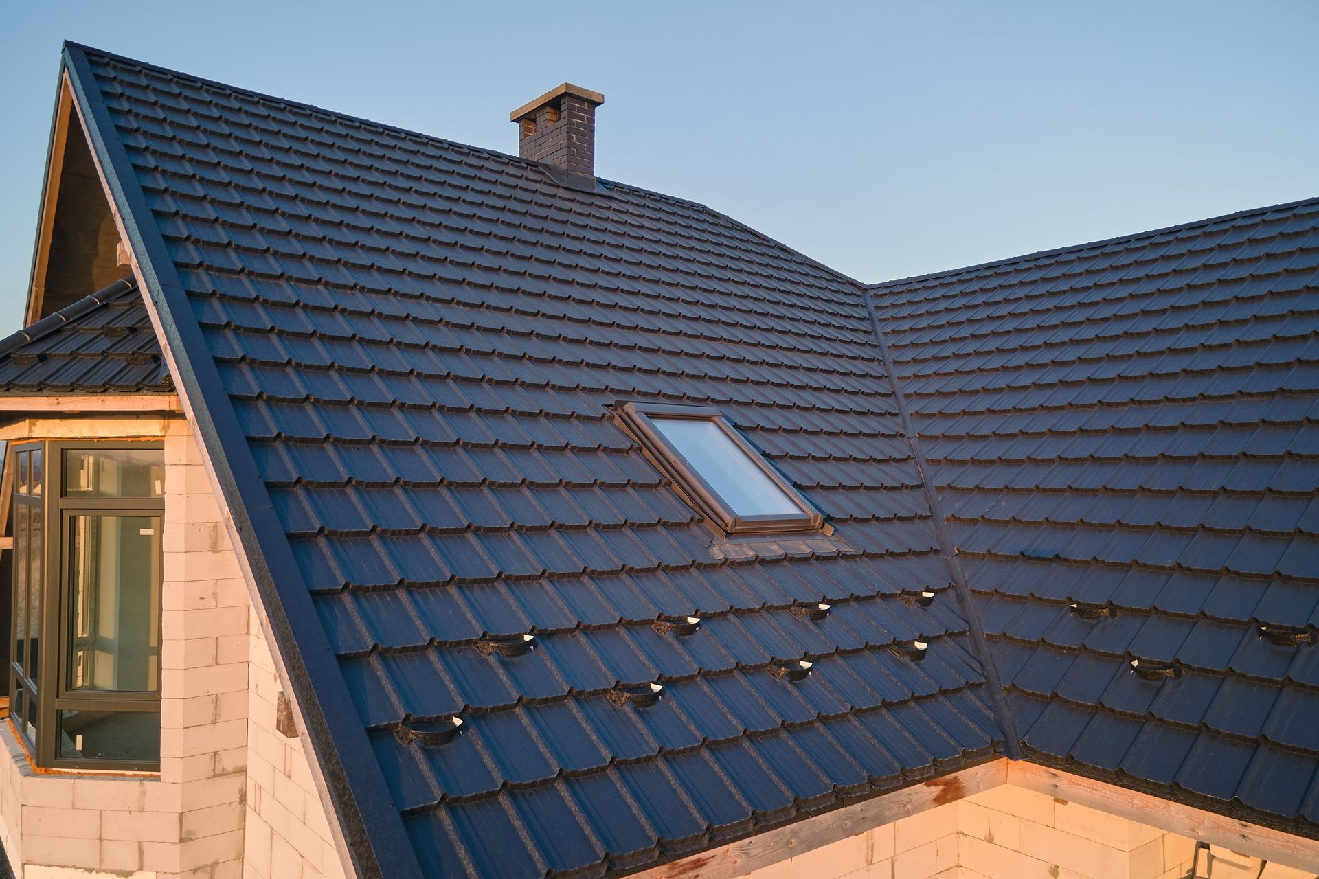 A close-up view of a dark, tiled residential roof with a chimney and a single skylight, set against a clear blue sky.