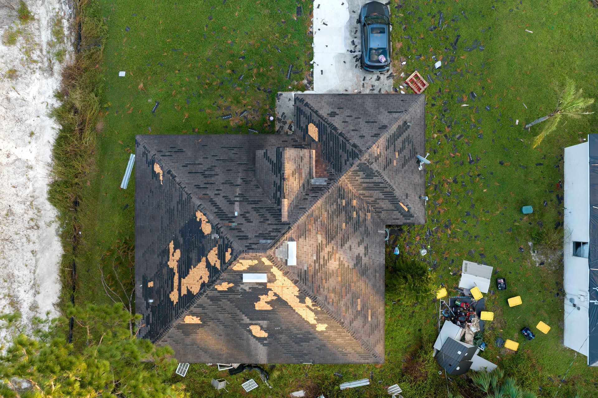 An aerial view shows a damaged residential roof with missing shingles, surrounded by debris scattered on the lawn.