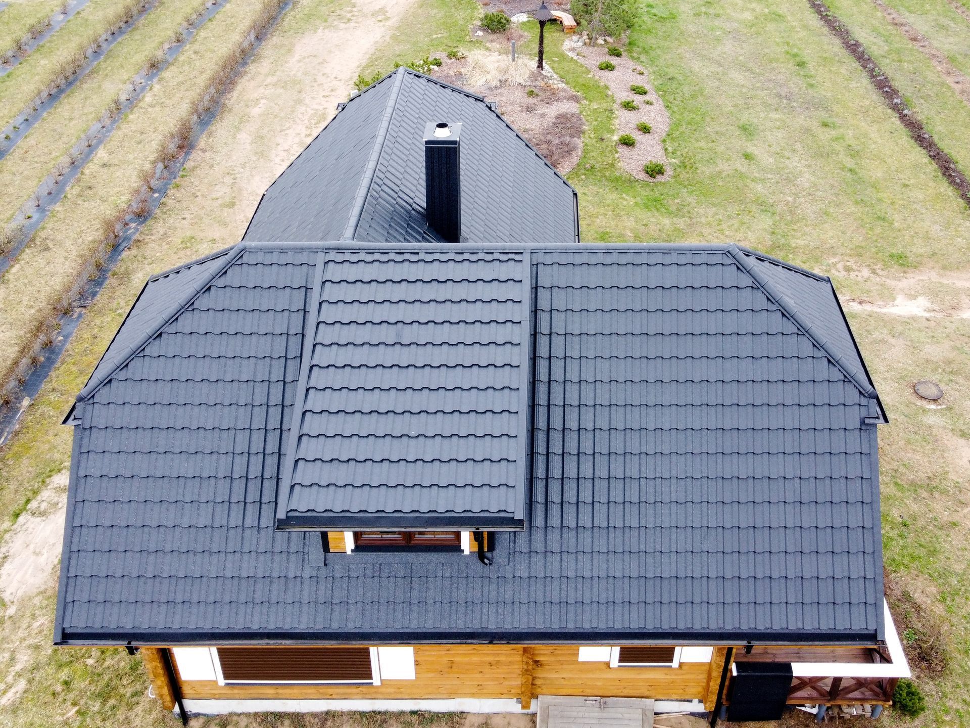 A high-angle view of a wooden house with a black metal tile roof and a prominent dormer window, surrounded by fields.
