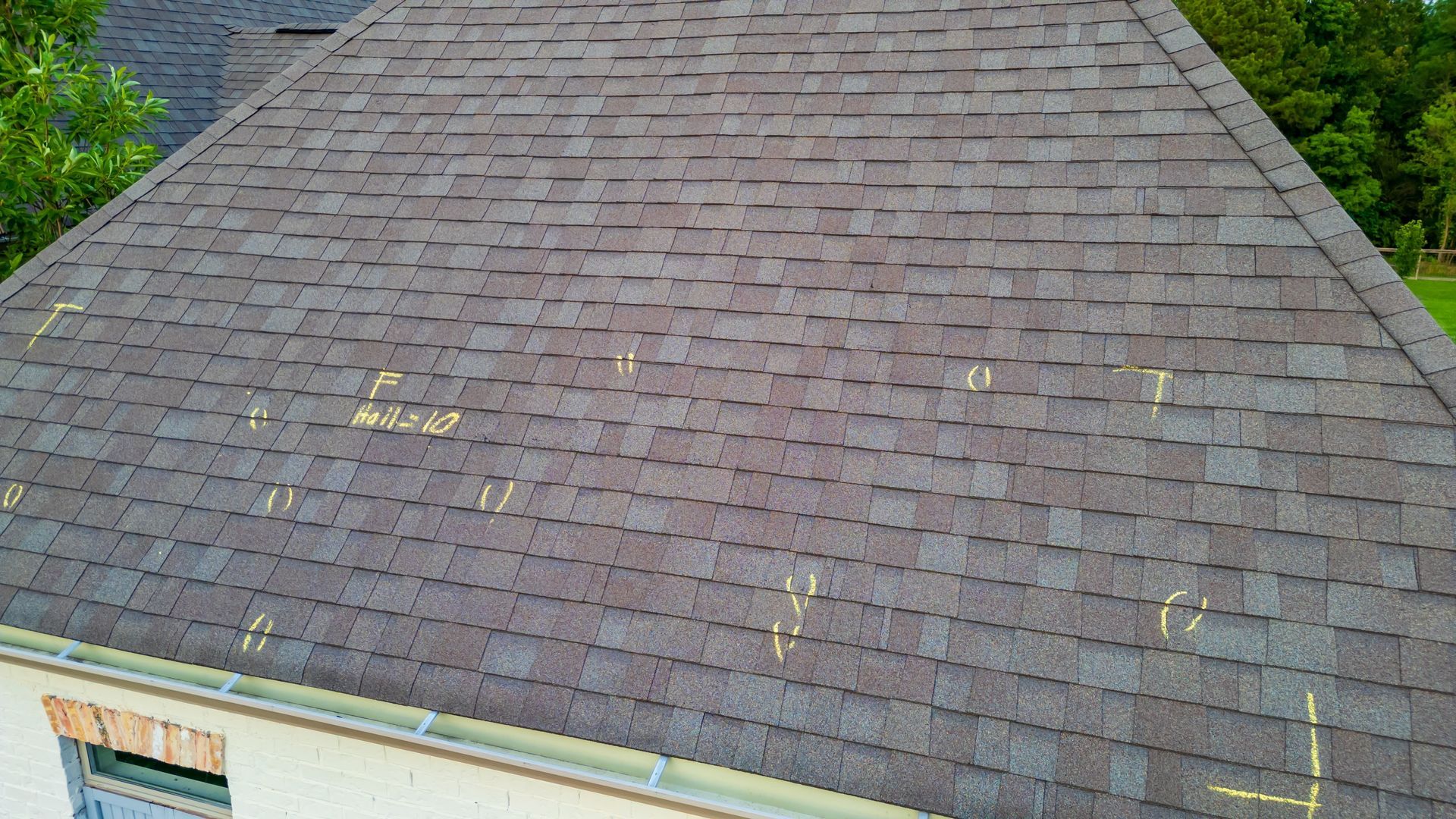 An aerial view of a dark shingled roof marked with yellow chalk circles and numbers, likely for hail damage assessment.