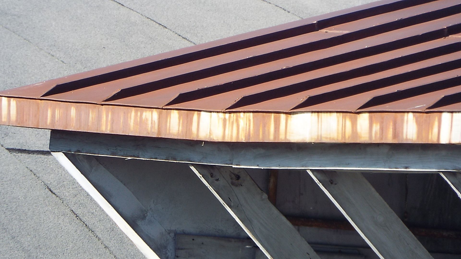 An angled close-up of a copper-colored roof with prominent ribs and a honeycomb-style edge, supported by metal beams.
