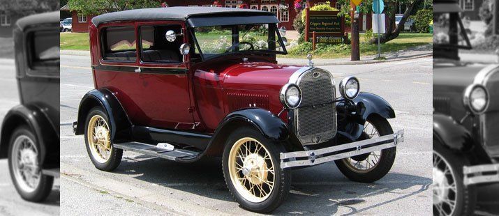 A burgundy 1930s Ford Model A car with black fenders parked on a street with other vehicles nearby.