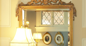 Ornate gold-framed mirror reflecting a window with diamond panes and two framed portraits.