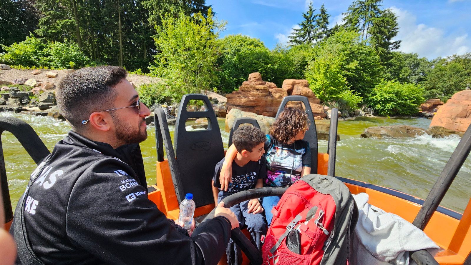 גבר וילדים בשייט. הסירה הכתומה על המים ליד סלעים ועצים.