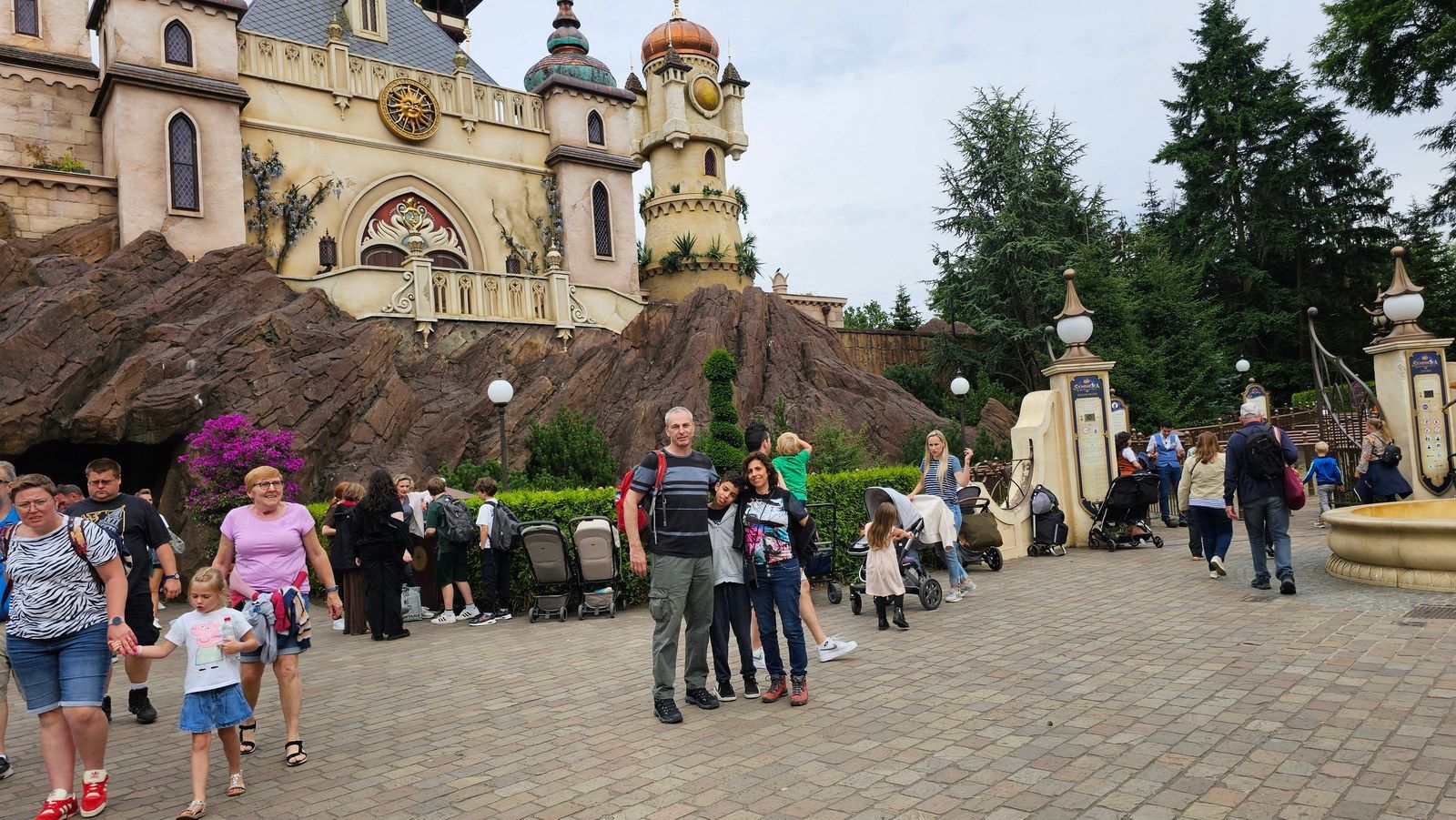 אנשים הולכים ליד טירה בנושא נושא עם מגדל שעון ומזרקה בפארק.